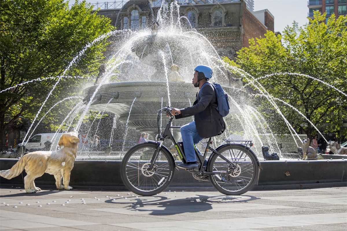 Greenworks e-bike — commuter riding past a city fountain with a dog